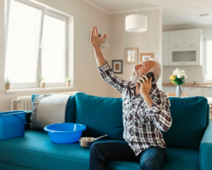 Older man, sitting on couch next to bucket that is collection water from leaking roof. Man is on phone, looking up at his roof, with hand in the air in frustration.